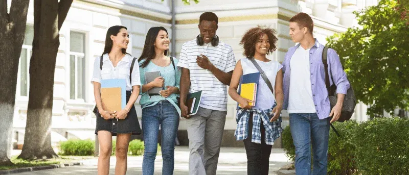 A diverse group of five smiling college students walk together on a campus path, carrying books and backpacks.